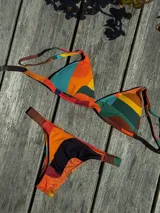 Conjunto de bikini con estampado geométrico multicolor en tonos naranja, verde, amarillo y marrón. El top es de estilo triangular con breteles regulables y cierre de gancho en la espalda. La bombacha es de corte vedetina con laterales elásticos fruncidos.