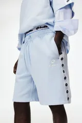 Short de felpa celeste de corte regular fit, con cintura elástica y cordón ajustable. Cuenta con bandas laterales blancas con el logo de Lotto repetido, bolsillos laterales y el logo de la marca bordado en una pierna.