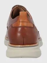 Zapato de vestir Hush Puppies modelo Rustic, confeccionado en cuero color marrón con cordones al tono. Presenta una suela de EVA en color gris claro con detalle de marca FLX en el lateral.