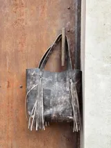 Bolso tipo tote de cuero con efecto desgastado, con dos bolsillos frontales y flecos largos decorativos a los costados. Cuenta con doble asa de hombro.