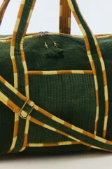 Bolso de viaje tipo duffel de color gris oscuro, confeccionado en algodón acolchado con pespuntes verticales. Cuenta con asas de mano y una correa larga regulable con detalles de tiras en tonos fucsia y naranja, cierre superior y herrajes metálicos dorados.