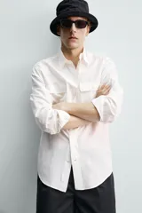 Camisa de corte relajado color celeste, confeccionada en mezcla de algodón y seda. Presenta cuello solapa, cierre frontal de botones, manga larga con puños abotonados y dos bolsillos de plastrón con solapa en el pecho.