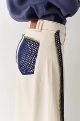 Jean de corte wide leg en color crudo, con detalle de franjas laterales tejidas en crochet azul y verde. Presenta costuras en contraste y ruedo deshilachado.