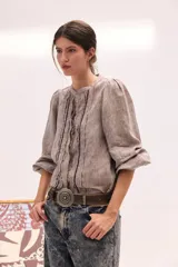 Blusa de lino color beige, de cuello redondo y manga larga abullonada. Cuenta con detalles de pliegues y ribetes oscuros en la pechera.