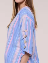 Blusa Erika de algodón con estampado de rayas en tonos azules, rosados y amarillos. Presenta cuello redondo con escote en V y botones frontales, mangas cortas decoradas con una fila de botones en los laterales y dobladillo asimétrico.