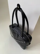 Cartera rígida negra con detalles en rojo y tachas plateadas. Tiene herrajes y cierre metálico plateado marca YKK, asa fina para llevar al hombro o en la mano y correa larga extra regulable. El interior es forrado, tiene un bolsillo con cierre enmarcado en cuero, y un segundo bolsillo plaqué.