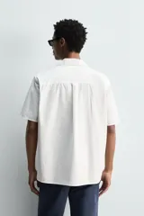 Camisa de manga corta con cuello bowling, confeccionada en popelín de algodón. Presenta un corte relajado, cierre frontal con botones y aberturas laterales en el bajo.