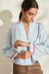 Blusa de lino color celeste con escote en V, cuello con volados y mangas largas con terminación de volados en los puños. Presenta detalles de vivos y costuras en tono rojo a contratono en los hombros y puños.