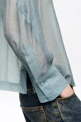 Camisa de corte boxy en color celeste, confeccionada en tejido semitransparente con apliques de pequeñas piedras brillantes. Posee cuello camisero, cierre frontal con botones y mangas largas con puños con sardineta.