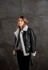 Campera tipo gamulán de eco cuero negro con interior y solapas de corderito sintético en color crudo. Presenta cierre frontal, bolsillos laterales con cierre y cinturón ajustable en la cintura.