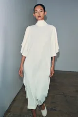 Vestido midi fluido de cuello alto y manga corta tipo capa. Presenta un diseño minimalista con bajo recto y aberturas laterales, con cierre en la espalda.