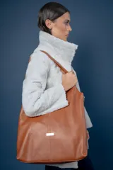 Bolso tipo shopper de gran capacidad, confeccionado artesanalmente en cuero vacuno color suela. Presenta un diseño minimalista con costura central vertical, placa metálica decorativa y fuelles laterales con cierre para ajustar su volumen. Incluye un sobre de cuero desmontable en su interior.