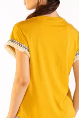 Remera de manga corta color mostaza, confeccionada en 100% algodón, con cuello redondo y detalles de flecos y guardas geométricas en las mangas.