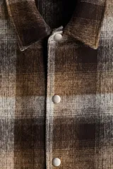 Sobrecamisa acolchada de corte relajado con diseño a cuadros en tonos marrones, grises y negros. Presenta cuello inglés, cierre frontal con botones a presión, hombros caídos y mangas largas con puños abotonados.
