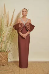 Vestido largo color marrón de corte recto, con escote profundo adornado con pedrería brillante. Presenta un diseño de hombros descubiertos con mangas voluminosas tipo lazo o drapeado, confeccionado en tela satinada.