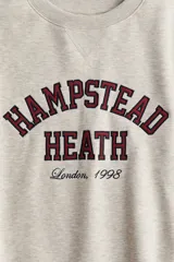Buzo oversized color beige con interior cepillado suave. Presenta hombros caídos y un diseño de texto en el pecho que dice "HAMPSTEAD HEATH London, 1998" en rojo y negro. Tiene ribete acanalado en cuello, puños y bajo.