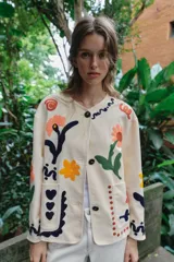 Chaqueta de algodón color crudo con cuello redondo, manga larga y bolsillos delanteros de plastrón. Presenta un diseño con bordados coloridos de flores y motivos abstractos a contraste, y cierre frontal con botones oscuros.