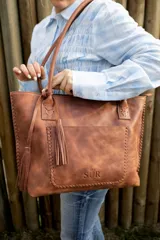 Cartera tipo tote de cuero marrón con costuras artesanales visibles, bolsillo frontal con el logo grabado, asas de hombro y detalles de flecos colgantes.