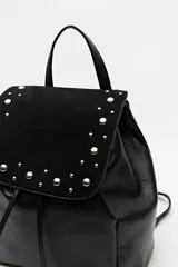 Mochila negra de símil cuero texturizado con tapa en acabado gamuzado, decorada con apliques de tachas metálicas. Tiene asa superior y correas regulables.
