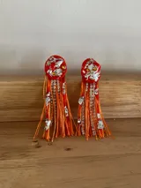 Caravanas artesanales de diseño llamativo, compuestas por una base circular de color naranja con apliques plateados y cristales, de la cual cuelgan flecos de hilo naranja y tiras de strass brillante.