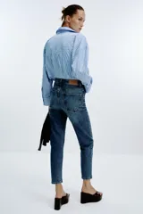 Jeans de mujer, tiro alto, corte Mom Fit con pierna slim y largo tobillero. Denim azul medio con efecto desgastado y lavado. Presenta cinco bolsillos y cierre frontal con cremallera y botón.