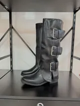 Botas de caña alta confeccionadas en ecocuero negro, con diseño de tres hebillas metálicas laterales y suela plana.