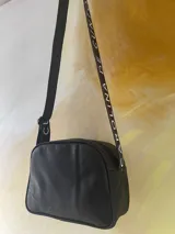 Bandolera de cuero negro y plateado con arte pintado a mano, perteneciente a la línea Ottenta de Carolina De Cunto. Cuenta con bolsillo frontal exterior, cierre superior con cremallera, forro interior con bolsillo y correa regulable con el logo de la marca.