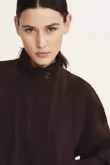 Campera corta oversize de color marrón, confeccionada en tejido de lino. Presenta cuello elevado, cierre frontal oculto con solapa y botones visibles, mangas raglán con puños abotonados, bolsillos delanteros con solapa y cordón ajustable en el bajo.