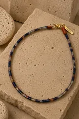 Gargantilla de cuentas tejidas con patrón de camuflaje en tonos azul, blanco y cobre. Cuenta con cierre de mosquetón metálico dorado y una pequeña placa con logo grabado.