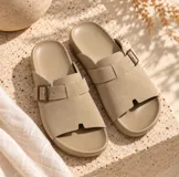 Sandalias planas de color beige con diseño de tira ancha en el empeine, hebilla metálica ajustable lateral y plantilla anatómica.