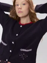 Cardigan de algodón grueso en color rojo, con cuello redondo y cierre frontal de botones grandes. Presenta bolsillos cuadrados en el bajo con detalles de pelo sintético en los bordes y logo bordado en uno de ellos. Posee puños y ruedo elastizados con diseño holgado.