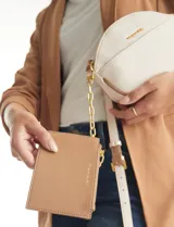 Cartera tipo bandolera de eco cuero con silueta de media luna, color beige y detalles en marrón claro. Incluye un monedero/tarjetero independiente en el bolsillo frontal y una cadena decorativa de eslabones dorados.