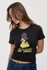 Remera blanca de manga corta, con estampa frontal de un pato de hule con turbante y labios pintados, acompañada de la frase "In all else fails, denial".