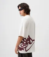 Remera oversized de algodón color blanco, de corte amplio y descontracturado, con cuello redondo, mangas cortas y gran estampa frontal localizada de inspiración urbana en tono bordó.