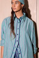 Camisa de corte holgado y relajado, confeccionada en popelina de algodón con diseño de rayas verticales en tonos verde, blanco y azul. Presenta cuello clásico, cierre frontal con botones y un detalle de cinta decorativa en zigzag de color azul marino a lo largo de la botonadura.