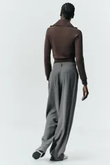 Pantalón gris de tiro alto con bolsillos delanteros y traseros, detalle de pliegues frontales y cierre frontal con cremallera y botón.