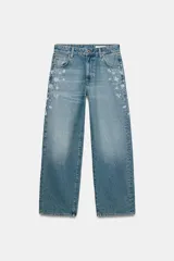 Jeans tiro medio de corte relaxed leg y largo completo, confeccionados en denim azul claro. Presentan detalles de aplicaciones de pedrería y joyas a contraste en los laterales superiores y bolsillos delanteros, estilo clásico de cinco bolsillos y cierre frontal con cremallera y botón metálico.