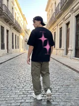Remera oversize negra con estampado trasero en color rosa vibrante. El estampado incluye dos figuras de estrella con efecto de brillo y texto en cursiva que dice "Limits are optional", "Rush the moment" y "Own the night".