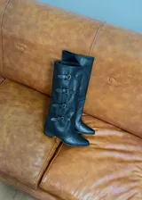 Bota de caña alta estilo slouchy color negro, con diseño de cuatro correas horizontales con hebillas metálicas doradas y punta redondeada.