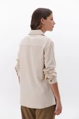 Camisa beige de manga larga, con cuello clásico, botones marrones y dos bolsillos frontales. Presenta costuras decorativas en contraste.