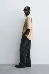 Camiseta de algodón color beige, corte relaxed fit, con cuello redondo y manga corta. Presenta un estampado gráfico grande en negro con motivos variados como una estación de servicio antigua, un caballo alado, una polilla, un pájaro y el texto "RUSTED ROSE".