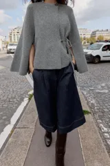 Chaqueta de punto gris con cuello redondo, manga larga acampanada y cierre frontal cruzado con botón y lazada lateral.