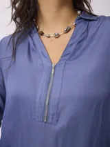 Blusa de satén color azul, con cuello camisero y cierre frontal parcial con cremallera. Presenta mangas largas con puños abotonados y un corte recto y fluido.
