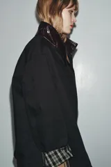 Chaqueta negra de la colección Zara Woman, confeccionada en hilatura con mezcla de algodón. Presenta un cuello alto brillante en contraste de color marrón oscuro, manga larga, bolsillos delanteros y pespuntes marcados. El forro interior es a contraste y el cierre frontal es con botones.