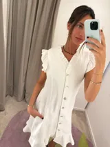 Vestido midi blanco con escote en V ribeteado con volados, frente abotonado y mangas cortas con volados. La falda es ligeramente evasé con un pequeño ruedo con volado.