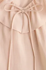 Blusa rosada de tejido liviano, con escote en V rodeado de volados y tiras finas para anudar. Presenta un diseño escalonado con volados anchos en el pecho y en el bajo, y mangas largas acampanadas con volados dobles en los puños.