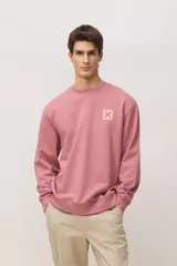 Buzo de cuello redondo en color rosado, con diseño de manga larga y puños acanalados. Presenta un pequeño parche cuadrado con una cruz en el pecho.