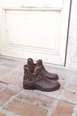 Bota corta de cuero color marrón, con taco bajo cuadrado y punta redondeada. Presenta un diseño estilo western con correas ajustables con hebillas metálicas y herrajes circulares en los laterales.