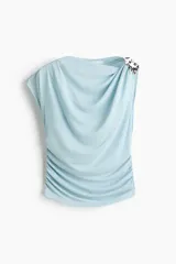 Top de punto celeste con leve brillo, sin mangas, con escote drapeado y detalle metálico martillado en un hombro. Presenta costuras fruncidas en los laterales para realzar el efecto drapeado.