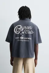 Camisa de algodón con estructura canvas, color rojo lavado, de corte relajado (relaxed fit) y manga corta. Posee cuello solapa, dos bolsillos de plastrón en el pecho y cierre frontal de botonadura. Presenta bordado y estampado de texto a contraste en el delantero.